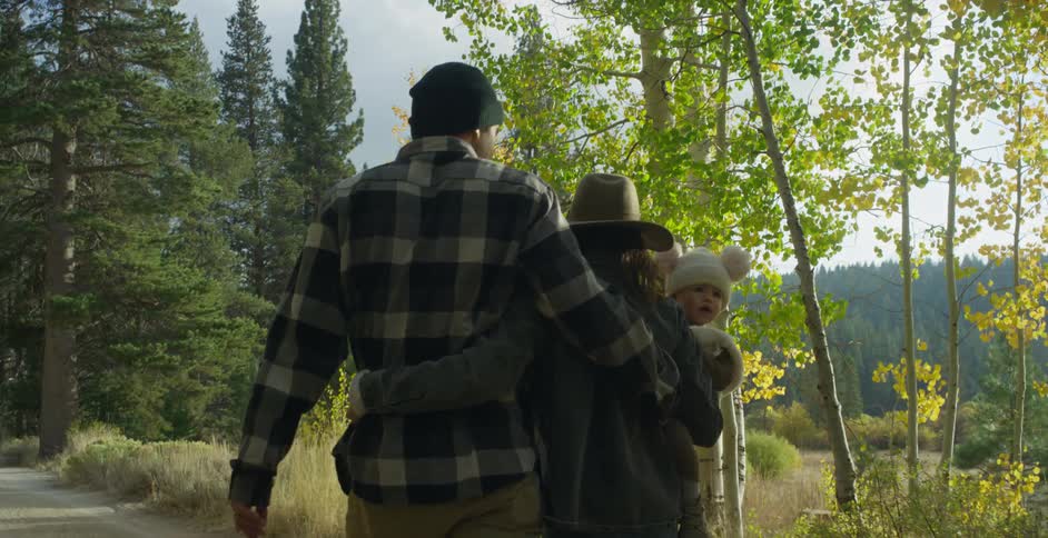 Family walks along dirt road across field from conifer forest on sunny day
