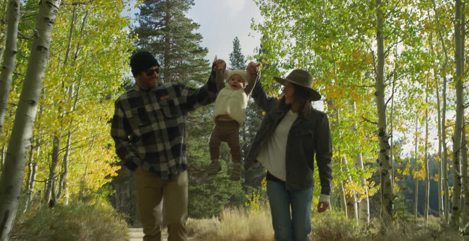 Mother with brown hat wavy hair and denim jacketand father wearing sunglasses green wooly hat and gray plaid shirt lift baby up walking in autumn forest
