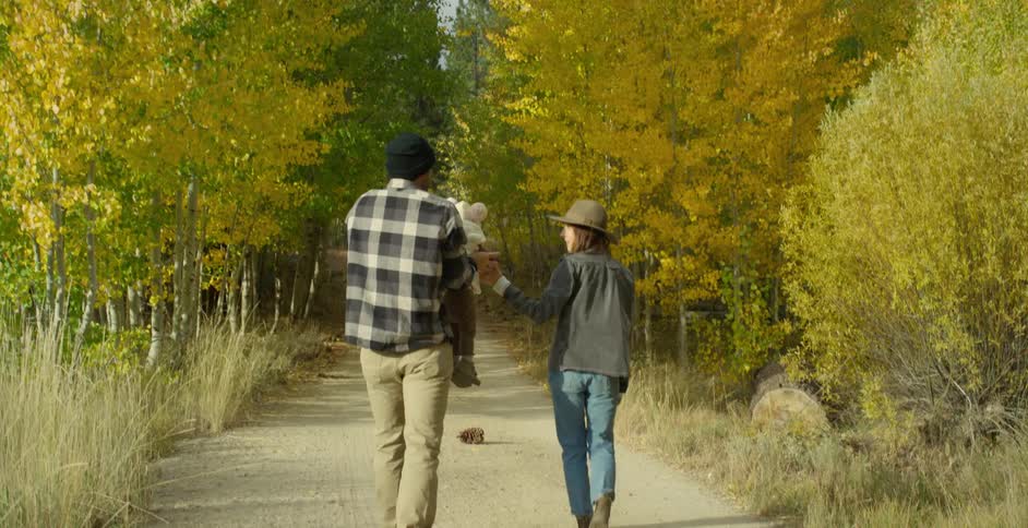 Father in green wool cap and gray plaid shirt and brown pants holds and carries toddler son next to mother with wavy hair in brown hat gray jacket and jeans in autumn forest