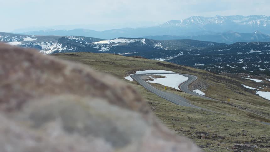 Snowy winding road on slope seen from hill behind large rock