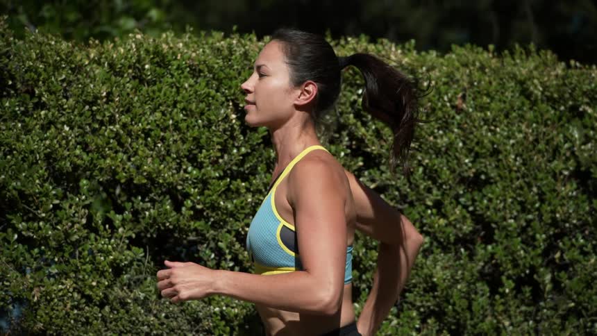 Woman in blue and yellow yoga bra jogging along hedge wall in slow motion
