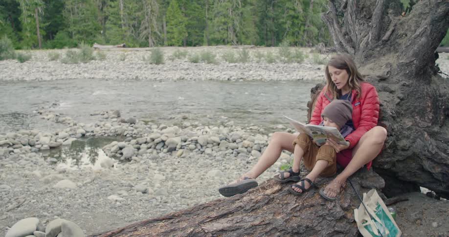 Mother and son sitting on a log near a flowing riverbed reading a book