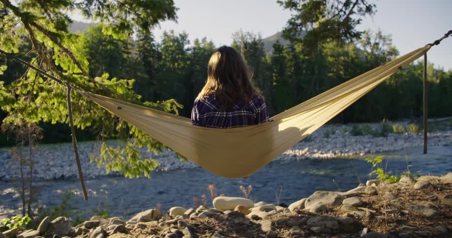 Woman with brown wavy hair and blue plaid shirt swings in hammock on river bank