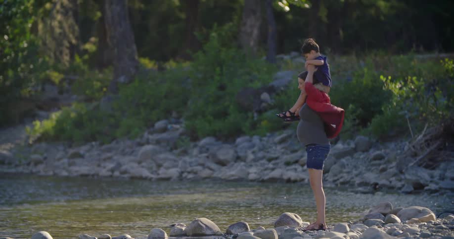 A pregnant mother carrying her son on her shoulders next to a river