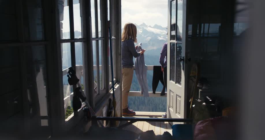Two women stand on a patio overlooking a mountain range and hang clothing on a railing