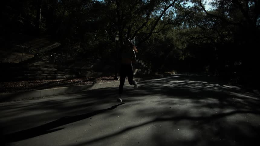 A female jogger runs along a road shrouded in shadow from the tall trees surrounding the road