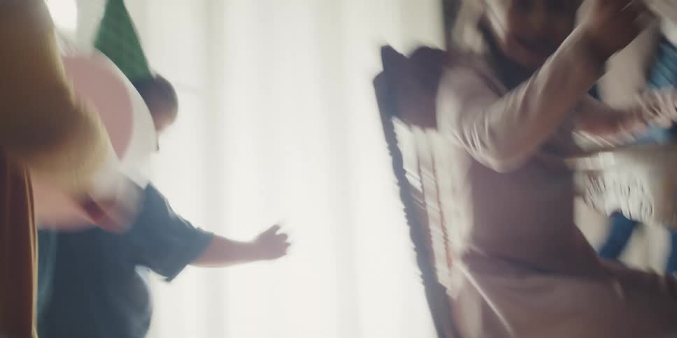 Children with party hats and balloons run through a house past children at a table and a woman