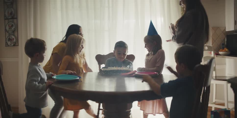 A young boy sits at a table at home and blows out candles on a cake and other children and two women clap