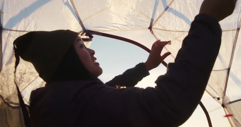 Woman wearing a warm coat and hat adjusts the straps in the interior of a tent on a sunny day