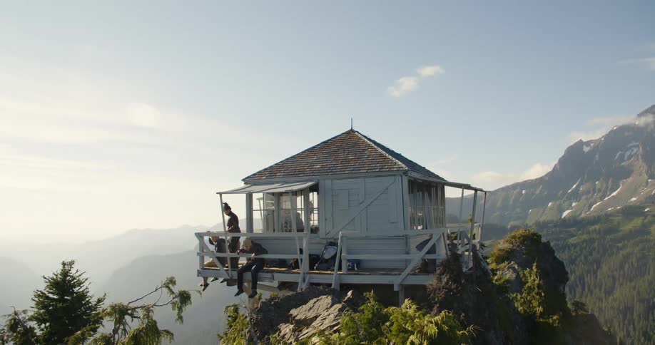 A small shack sits on a cliff and two people sit on the deck with their legs dangling over and another woman walks over and sits down with them
