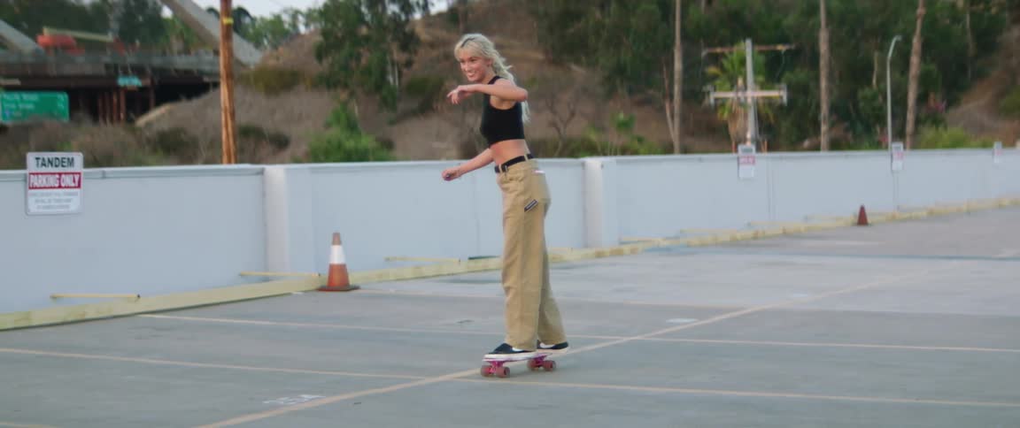 Group of friends filming a young woman learning how to ride a skateboard on a parking lot at sunset
