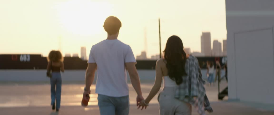 Man carrying a water bottle holds hands with a woman as they walk towards a group of people outdoors