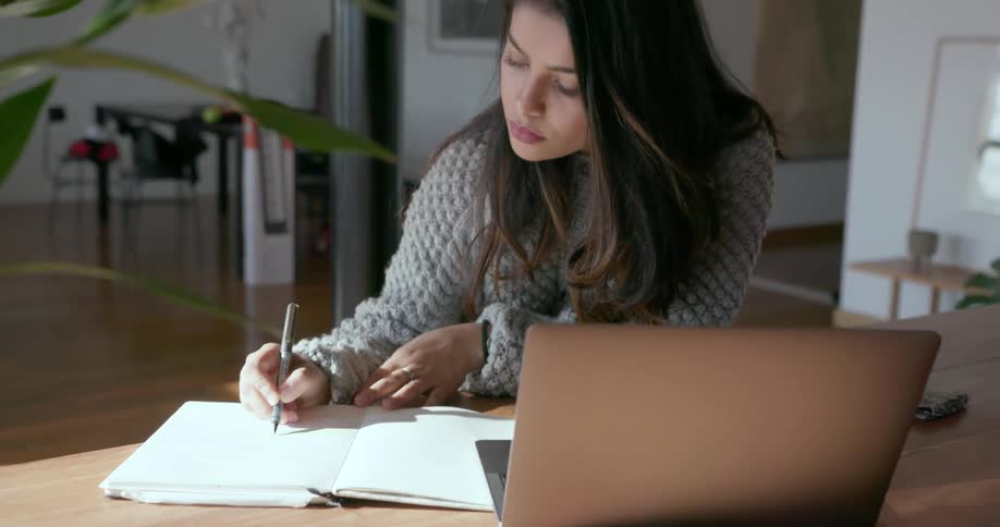 A woman sitting at a table with an open laptop makes notes