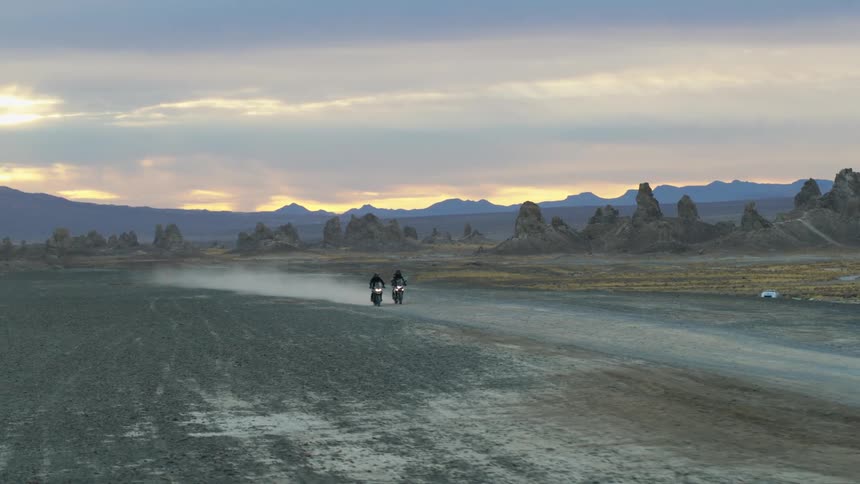 Two riders ride motorcycles and stir up dust on a dirt road near a field and mountains