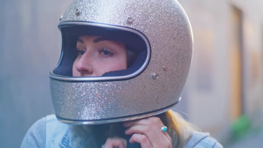 Woman with brown hair and blue jacket puts on silver motorcycle helmet