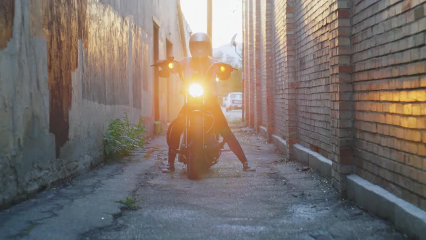 A woman wearing a silver glitter motorcycle helmet is on a motorcycle and she is parking in an alley between two buildings