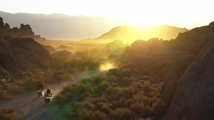 Two motorcycles stir up dust as they travel on a dirt road near the bushes on a sunny day