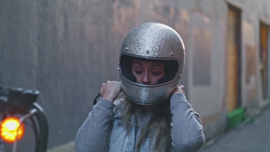 Woman with fair hair and denim jacket puts on silver motorcycle helmet in the street