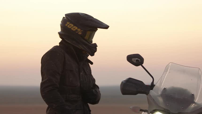 Male biker with helmet and black jacket stands next to motorcyle at dusk in the desert