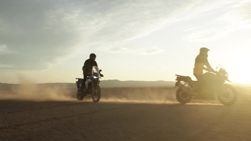 Two motorcycles stir up dust as one passes the other in the desert on a sunny day