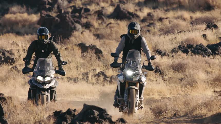 Two men ride motorcycles through heavy brush and boulders nestled between hills