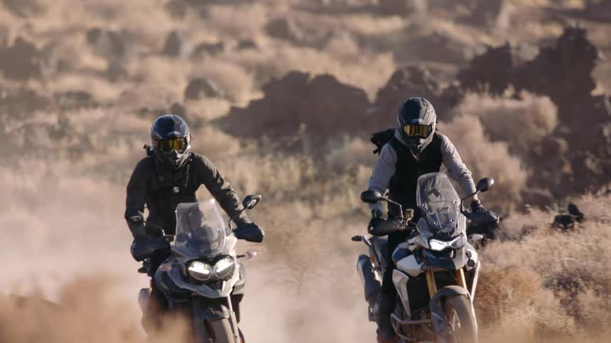 Male bikers with motorcycle helmets and black jackets ride motorcycles in the desert