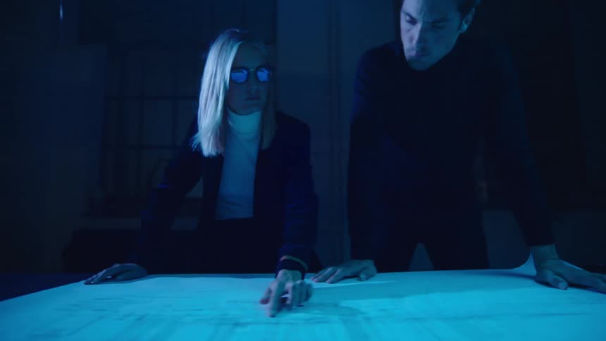 Woman wearing glasses slides her finger across a large diagram as a man stands next to her and watches