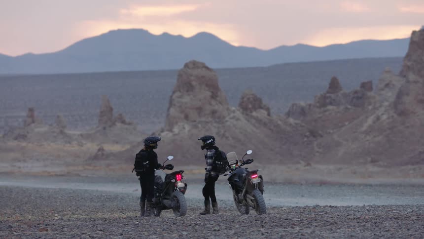 Bikers with black motorcycle helmets jackets pants and backpack get off bike in the desert at dusk