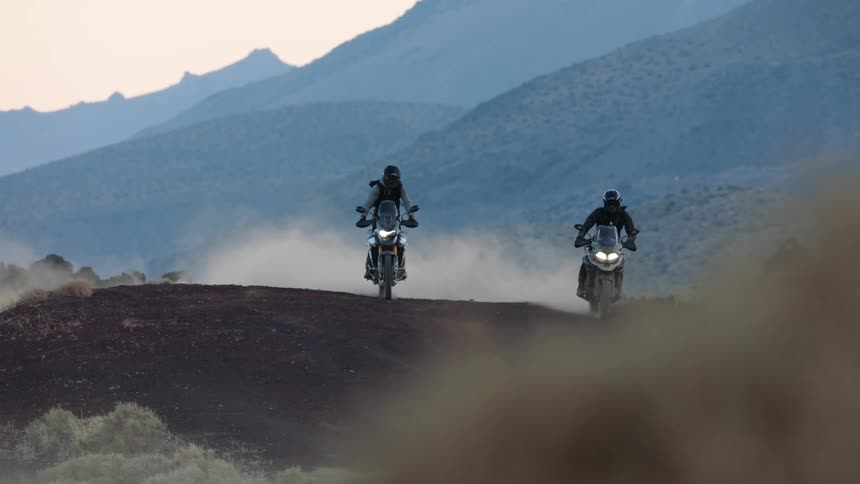 Two people kick up dirt as they ride motorcycles through rolling hills in a canyon