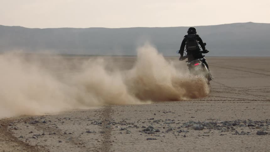 Biker with brown boots black pants jacket and backpack starts motorcycle and skids in the desert
