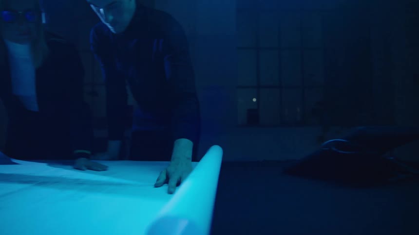 A man and woman roll out blueprints on a table in a dark room