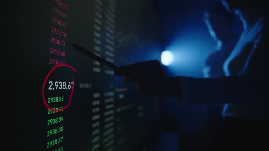 Man and woman discuss stock numbers on large digital display in dark room lit by single bright blue light