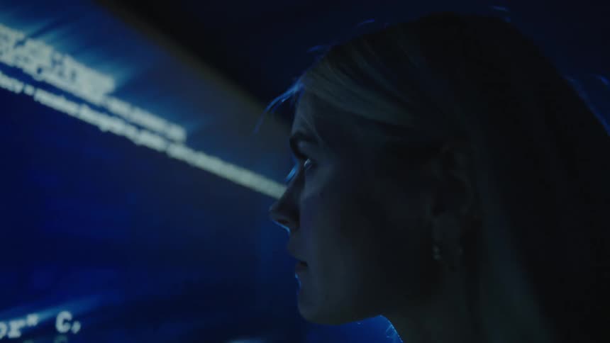 Woman with fair hair and white turtleneck sweater looks at big screen with numbers and graphics in dim lit room
