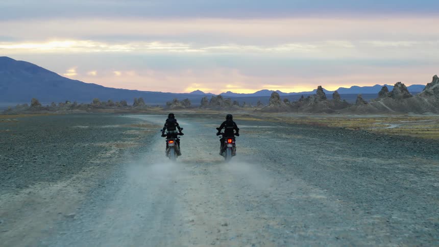 Two motorcyclists ride over gravel toward brightly lit sky over blue misty mountains