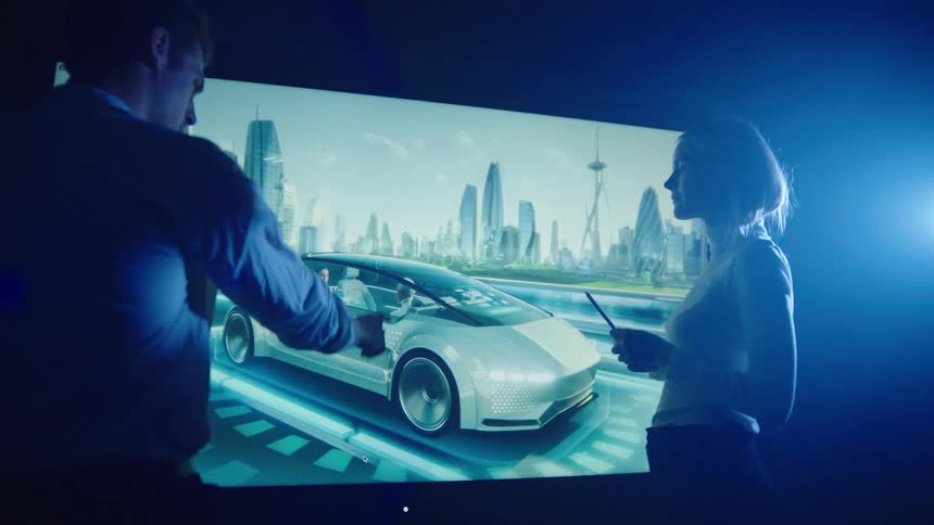 Man and woman standing in dark room lit by single light discuss image of car on highway in city that is displayed on a large digital screen