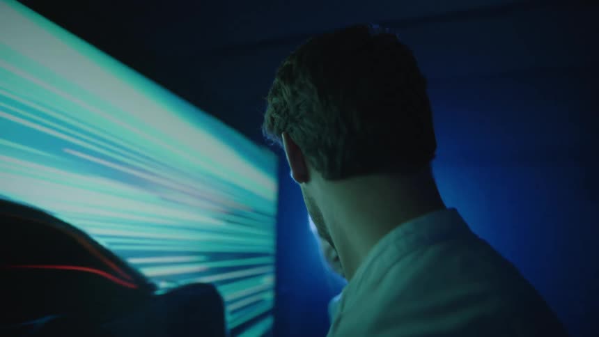 Man and woman stand in front of a large screen and smile as they watch a car traveling on the screen