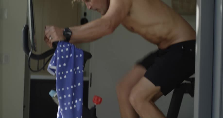 Shirtless blonde man with curly hair rides stationary bicycle