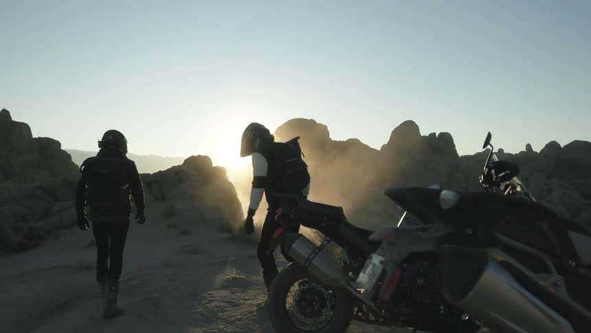 Two men get off their motorcycles and walk down a dirt road as the sun sets
