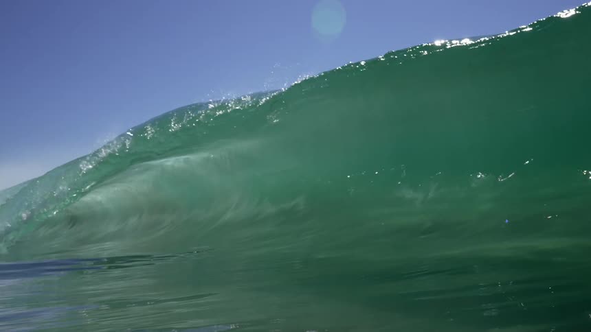 The sun shines brightly on a big blue wave as it rolls overhead