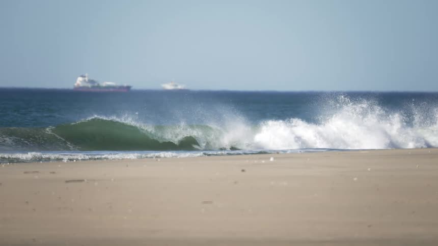 Waves crash on the sand of a beach next to the ocean with cargo freight ships on the horizon