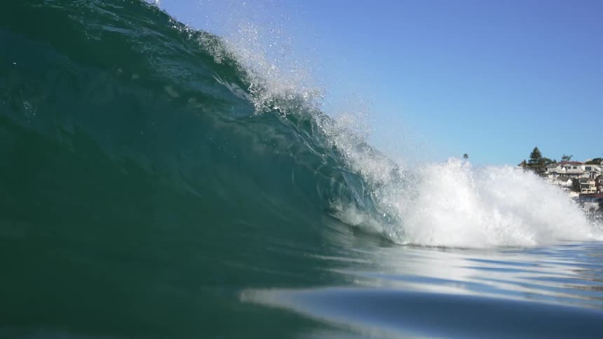 Large ocean wave rolls and splashes in front of the buildings on the coast under a blue sky