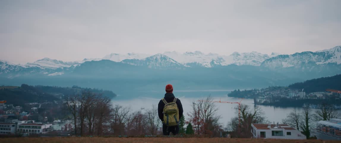 Male hiker wearing backpack stands on hill overlooking town lake and snow covered mountains