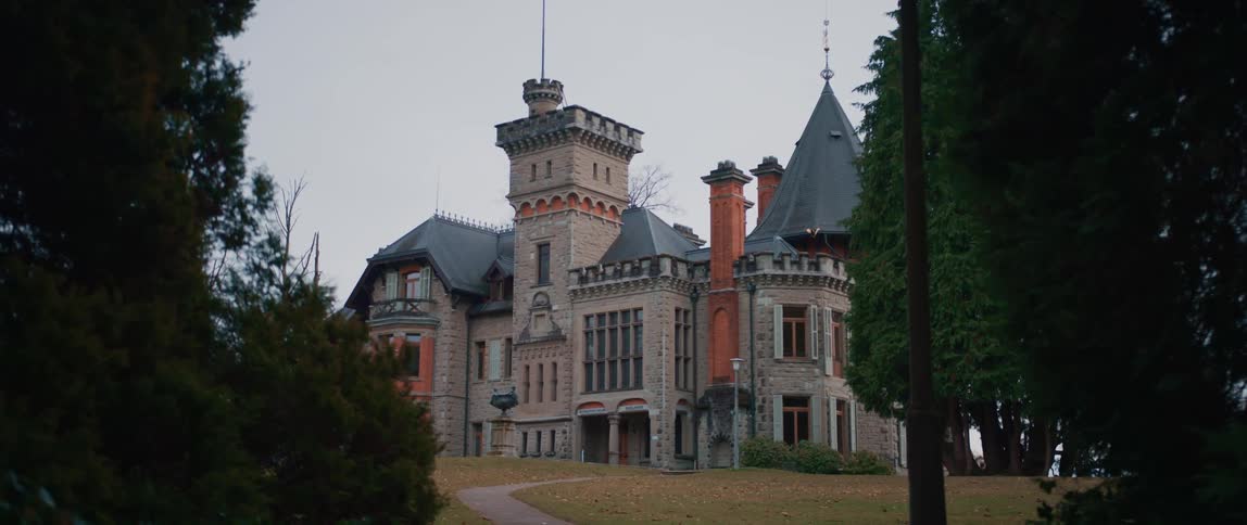 Large ornately decorated and architecturally detailed mansion is bordered by wide lawns and conifer trees