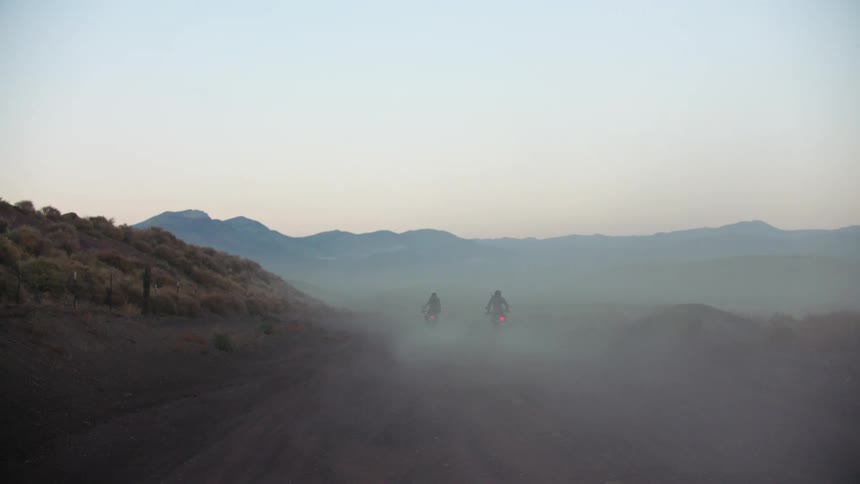 Two bikers with black motorcycle helmets jacket and pants ride motorcycles on country road at dusk