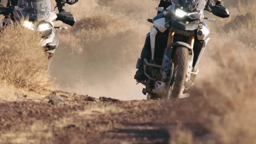 Two men ride motorcycles down a winding road through bushes
