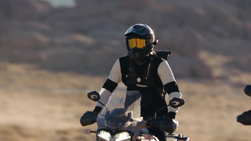 Two motorcyclists ride motorcycles on a dirt path near the bushes in the desert