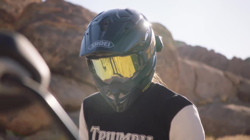 Woman standing near large boulders wearing helmet dons gloves and walks over to motorcycle
