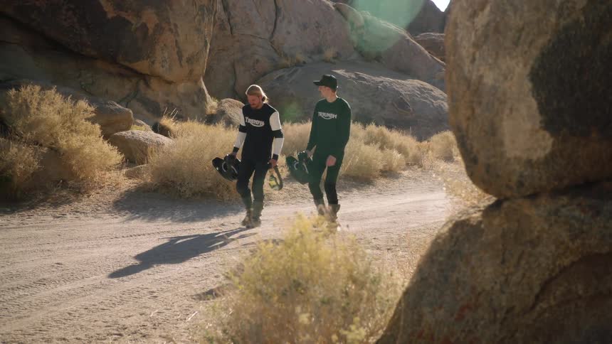 Two men carrying helmets walk along a dirt road through a canyon