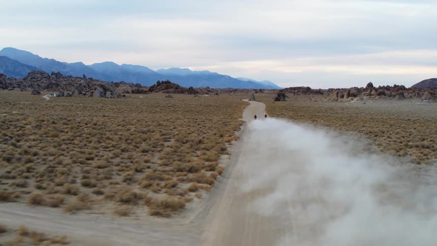 Two motorcyclists kick up dust as they ride down a dirt road in a field of bushes