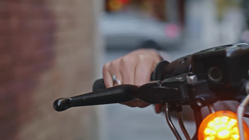 Woman sitting on cycle near brick wall revs handle as cars moves along street